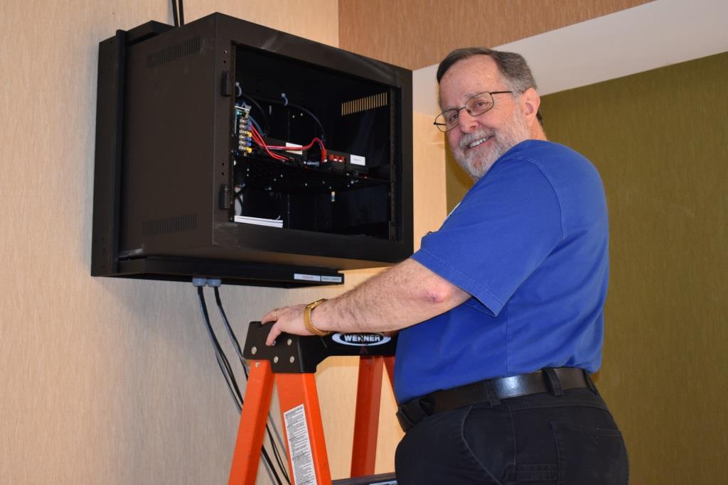 Paul, N3RQV, is standing on a stepladder next to the wall-mounted radio cabinet. The power supply and TM-D710 radios can be seen inside the cabinet.